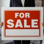 Close-up of a person in a suit holding a For Sale sign in a furnished room.